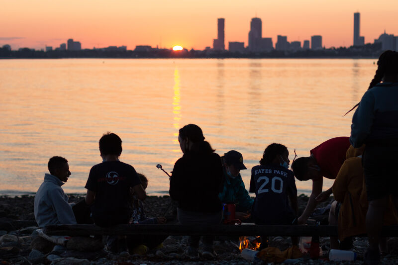 The image shows a group of people gathered around a fire on a beach at sunset. The sky is filled with warm colors, and the sun is setting over the water. In the background, there is a city skyline. The people are silhouetted against the light, and some of them are roasting marshmallows.